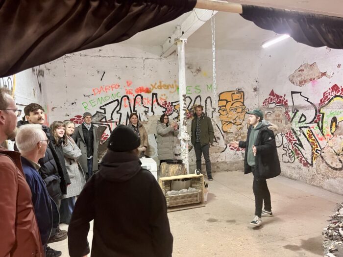 Man som står och talar framför en grupp människor. De befinner sig i ett rum målat med graffiti på väggen.